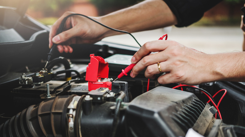 Hands test a battery under the hood of a car.
