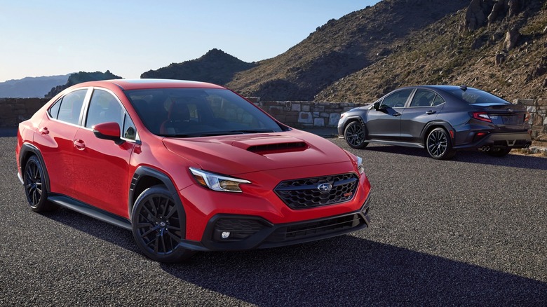 A red and a gray Subaru WRX in front of a hill.