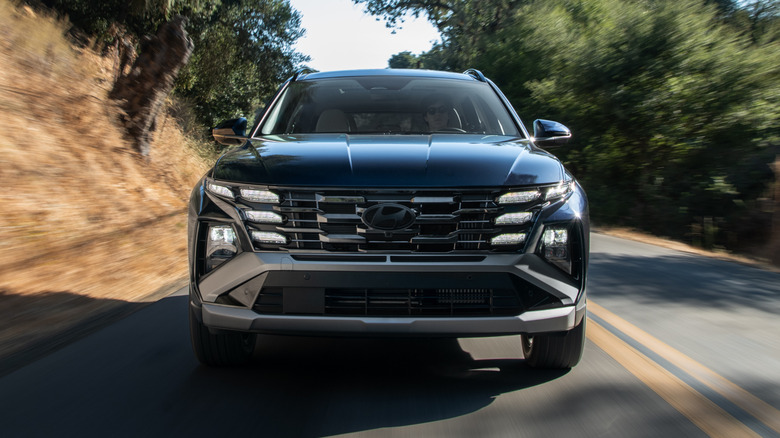 Front view of a black 2026 Hyundai Tucson Hybrid driving on a road surrounded by hills and trees
