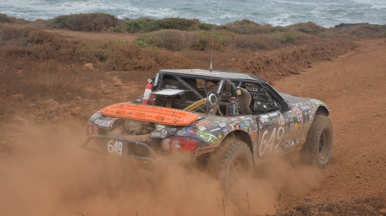 an off-roading miata covered in stickers drives towards the ocean while kicking up a cloud of dust