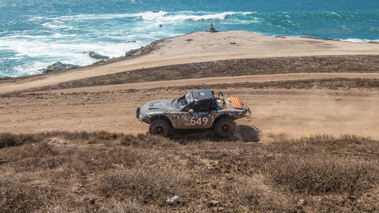 An off-road Miata covered in stickers and the number 649 painted on the side driving down a dirt road next to the ocean