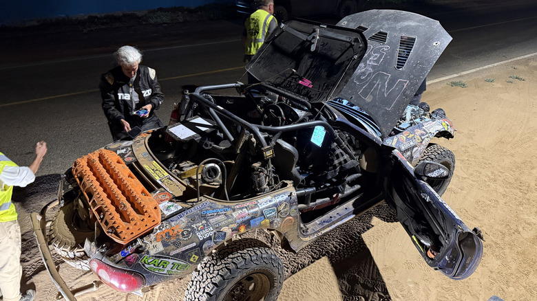 a spotlight illuminates an off-roading miata on a lift as people work on it at night