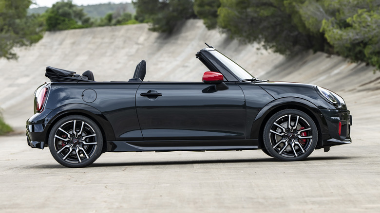 A side view of a black JCW Mini Cooper S Convertible parked with its top down on concrete in front of trees