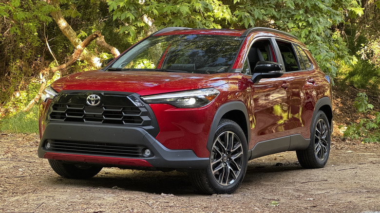 A front three-quarters shot of the red gas-only Corolla Cross parked on dirt in front of trees with its headlights on