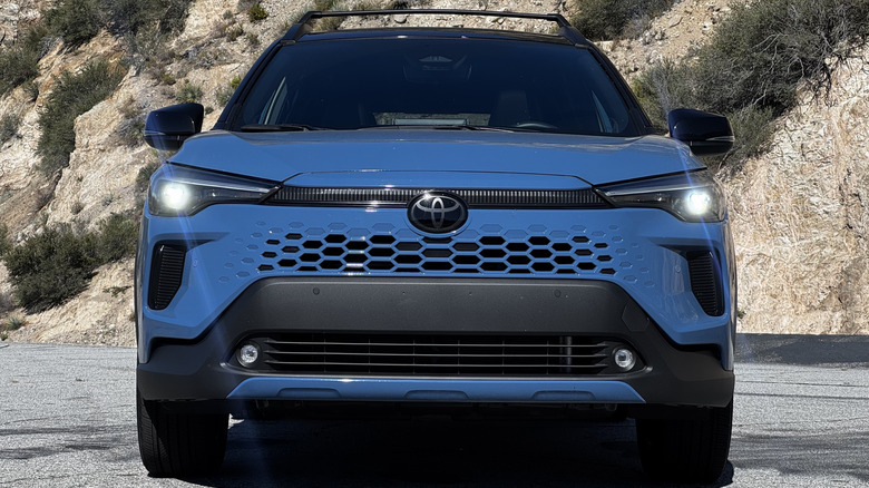 A head-on shot of the blue Corolla Cross Hybrid with its headlights on parked on asphalt in front of a shrub-covered mountainside