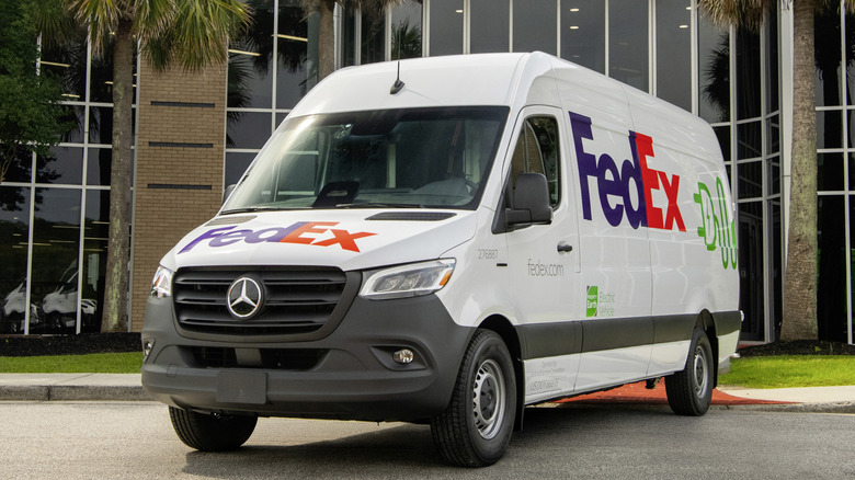 A front three quarter shot of a white Mercedes-Benz Sprinter EV panel van with a FedEx livery parked in front of palm trees and a building with bricks and glass