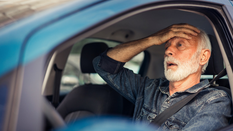 An elderly man behind the wheel of a car with his palm on his forehead