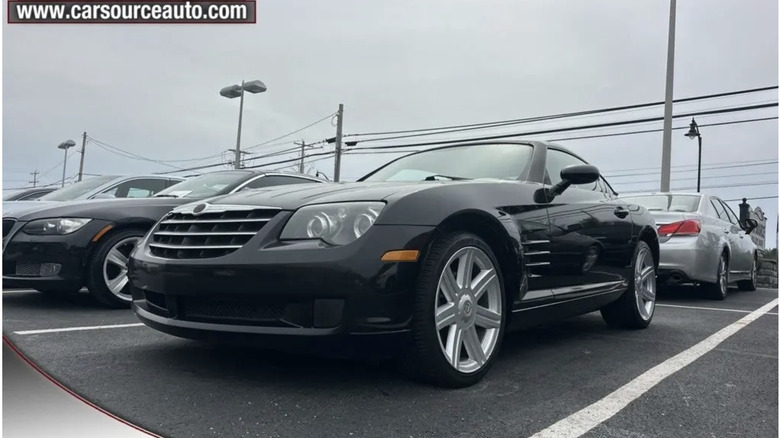 A black Chrysler Crossfire parked on a used car parking lot under a cloudy sky