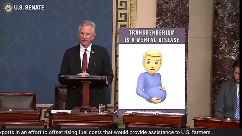 Tommy Tuberville, a real shitheel, stands by a sign with an emoji that he's very upset about