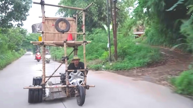 A double-decker cube extreme Vespa constructed of Bamboo and rubber ties with a wood panel backing the upper level. Each wheel position has three wheels except for the front driver side. It's driving along a roadway surrounded by plants and trees.