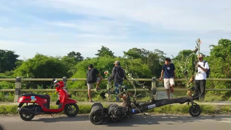 A red Vespa is parked behind an extreme Vespa which looks like a log with a wheel up front and two odd rows of wheels at the rear, on a bridge with four individuals looking out onto a river surrounded by trees.