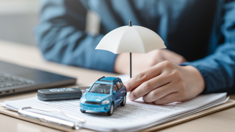 A person's hand holding a tiny umbrella over a blue car placed atop a stack of papers secured to a clipboard