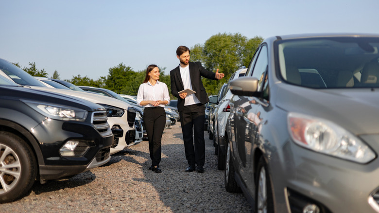 Man shows woman a car on a dealership parking lot.