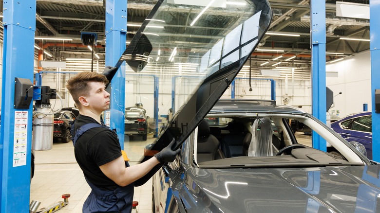 A repair tech carries a new windshield to a gray SUV in a collision repair center