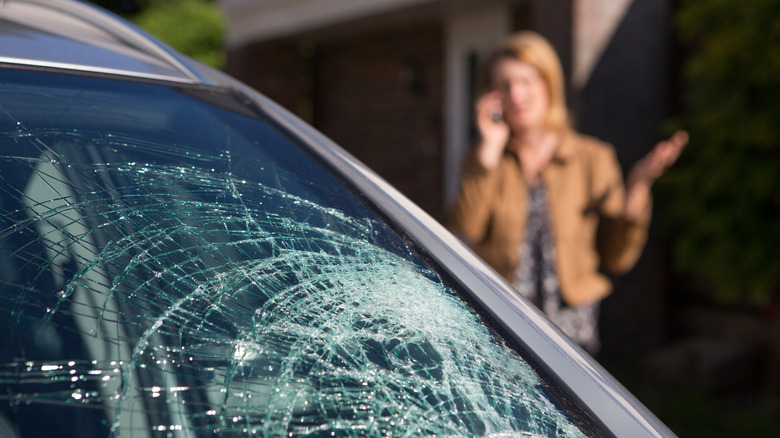 A blurry background woman in a brown coat calls someone to complain about the car with a broken windshield in the foreground