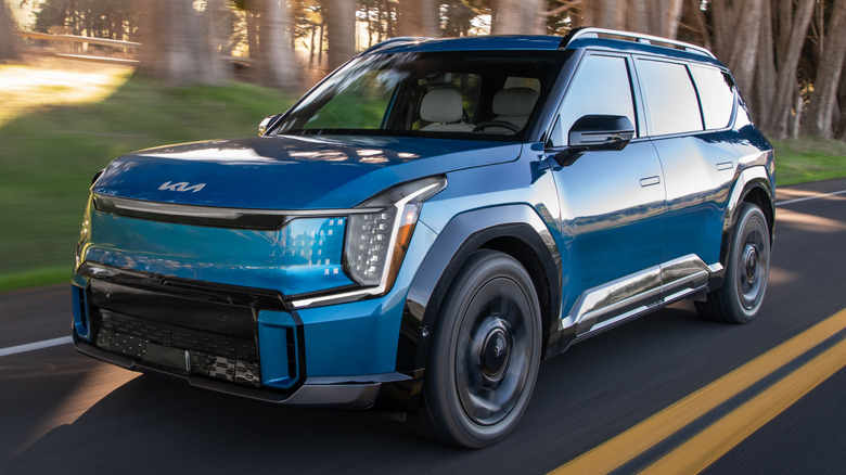 A 2025 Kia EV9 driving down a road with tall trees in the background