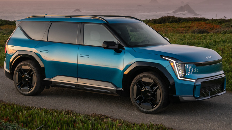 A blue 2025 Kia EV9 parked on a paved road along a coastline