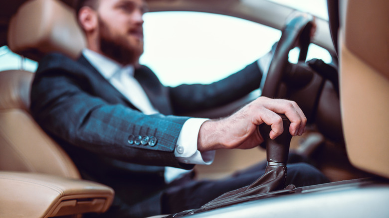 A man in a suit, driving a car with a manual transmission