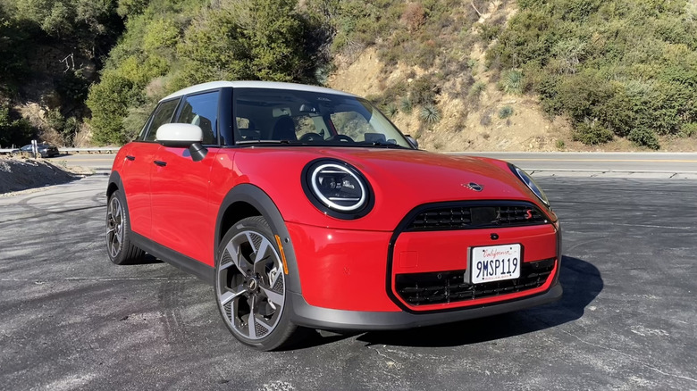 Front three quarters shot of a red Mini Cooper S four-door hardtop parked on blacktop in front of a shrub-covered mountainside