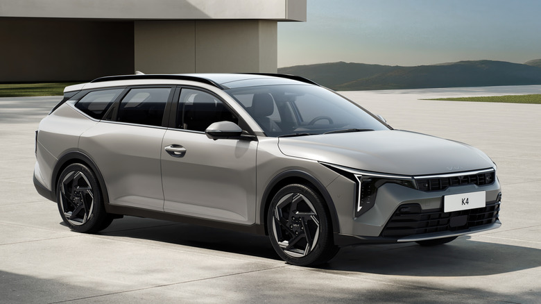 A front three quarters shot of a silver K4 Sportswagon parked on concrete in front of a concrete building and a mountain range