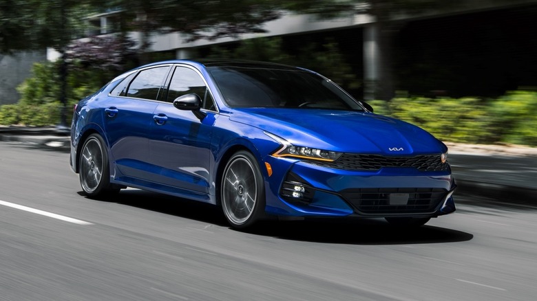 Blue 2024 Kia K5 driving down city street next to a building