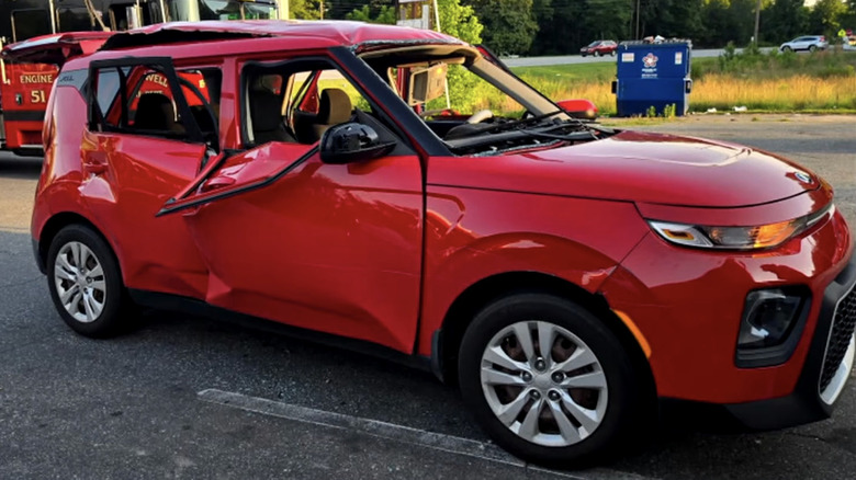 A damaged red Kia Soul that exploded from the inside. It has dents in the side, no window glass, and a popped-out roof