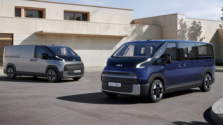 A front three-quarters shot of a dark blue PV7 passenger van parked next to a silver cargo van in front of a white modern home
