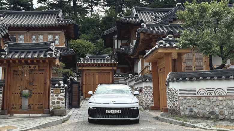 A head-on shot of the white Grandeur parked between two ancient traditional Korean homes in front of giant pine trees