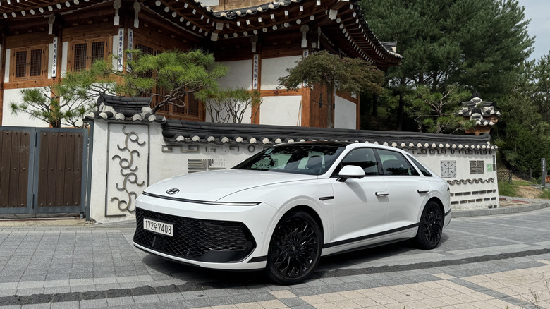 Front three-quarters shot of a white Grandeur parked on gray brick in front of a traditional ancient Korean house with black roof tiles