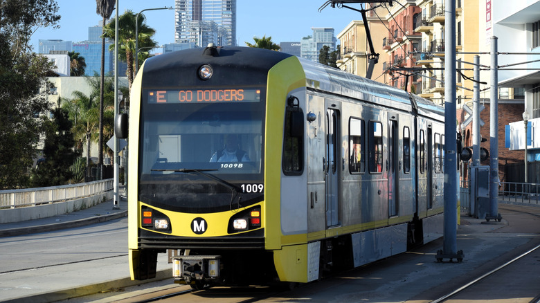 A photo of the LA Metro E-line train on the street passing apartments with a glimpse of a skyline behind it