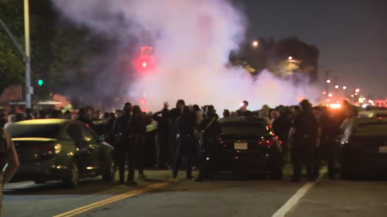 Smoke from an in-progress street takeover rises above a crowd of people and cars in an intersection as LAPD descends.