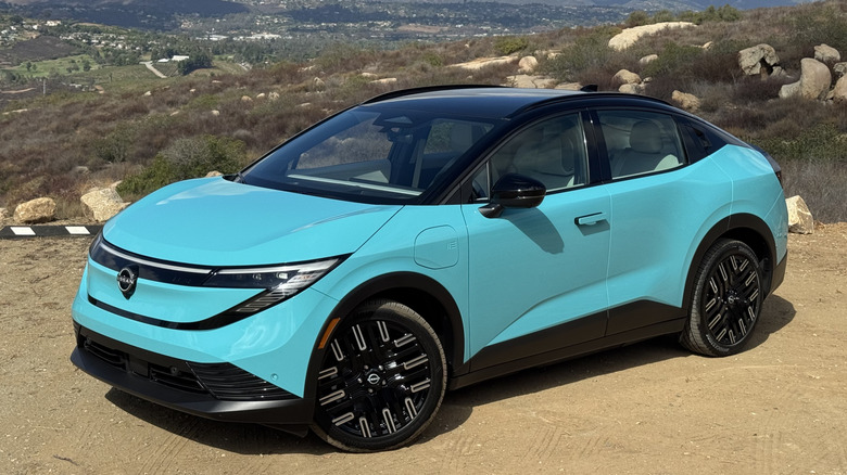 A front three quarters shot of a turquoise 2026 Nissan Leaf parked on dirt in front of rocks and shrubs and a valley