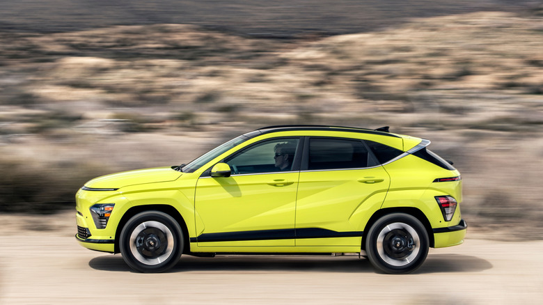 A side view of a highlighter yellow Kona Electric driving on a dirt road past a blurry desert landscape