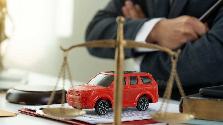 Judge sitting behind desk, holding gavel. Red toy car sits behind legal scales.