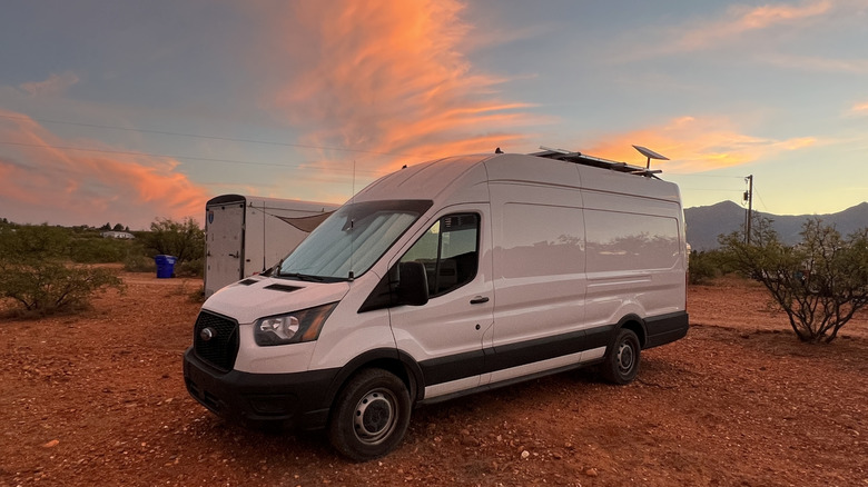 Front 3/4 view of a white Ford Transit camper van in the desert at sunset with orange clouds above
