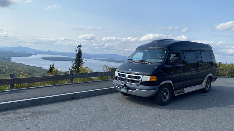 Front 3/4 view of a Dodge Ram conversion van with a lake, trees, and mountains in the background