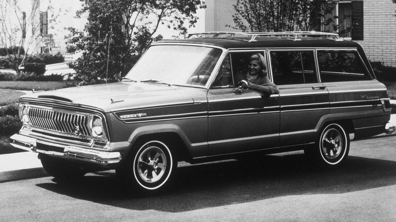A black-and-white photo of a 1966 Jeep Super Wagoneer in front of a house, with a woman leaning out of the driver's window.