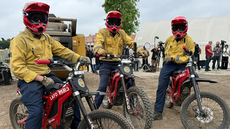A screenshot showing three firemen wearing yellow jackets and red helmets sitting on the red E-motorbikes in front of a bulldozer and reporters