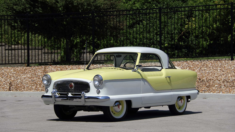 A yellow and white Nash Metropolitan parked in a sunny parking lot with a fence and trees in the background