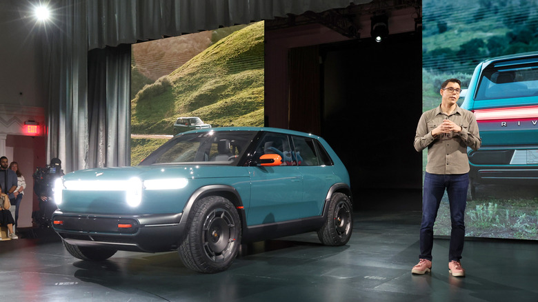 A blue-green Rivian R2 all-electric compact SUV parked on a stage surrounded by images of the car next to Rivian CEO rj scaringe