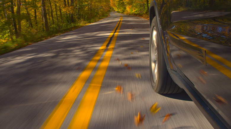 A car tire and driver-side door, driving down a two-lane road.