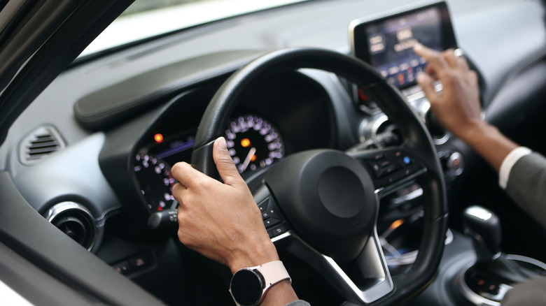 A person driving, with one hand on the wheel and the other extended to navigate the touchscreen controls.