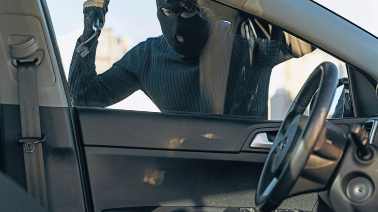 A masked man with a crowbar looking inside a car before breaking in