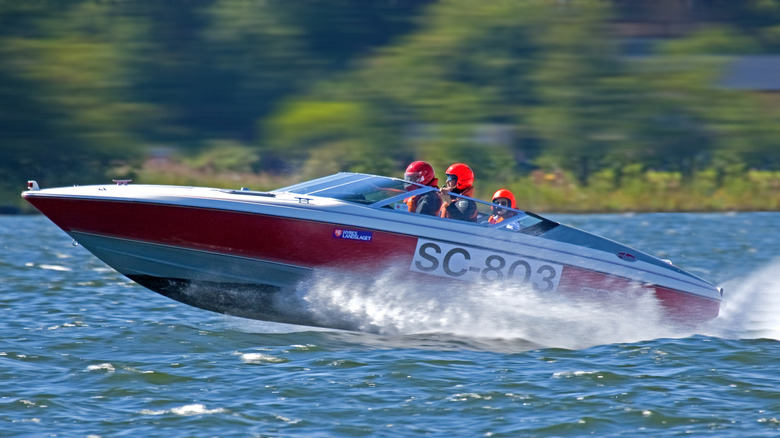 A red and white racing boat with three people in orange helmets speeds past blurred trees in background