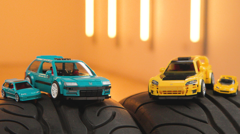 A teal Honda Civic EF Mattel Brick Shop car and Hot Wheels car sitting on a tire next to a yellow S2000 Brick Shop car and Hot Wheels car on another tire in an orange room