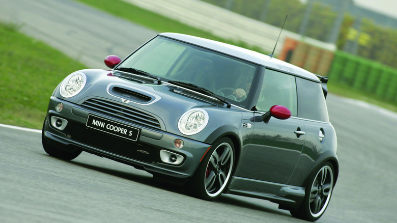 A front three quarters shot of a gray first-gen Mini Cooper S JCW driving on a race track next to green grass