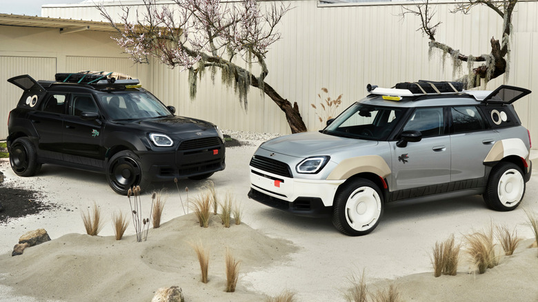 The two custom cars parked facing eachother at a front three-quarters angle in front of trees and a beige fence surrounded by mounds of sand and some plans