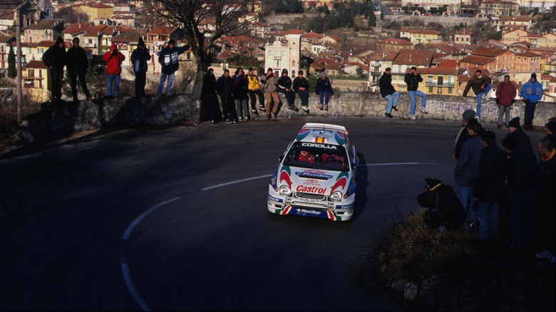 A Toyota Corolla WRC rally car rounding a hairpin turn lined with spectators above the town in the 1999 Monte Carlo Rally