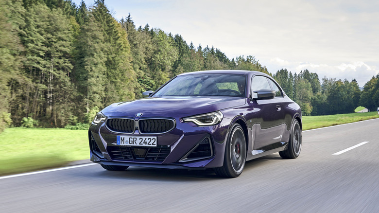 A purple BMW 2 Series coupe driving on a highway with a dense forest in the background