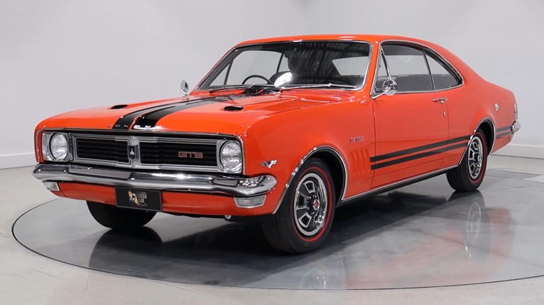 A vibrant orange Holden Monaro GTS coupe positioned on a reflective silver turntable inside a minimalist white studio.
